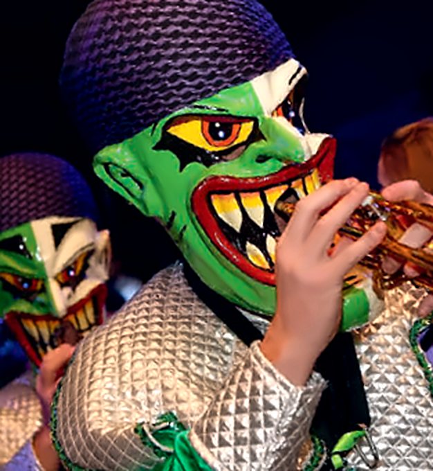 Grüne Maske mit gelben Augen und spitzen Zähnen beim Trompetespielen, dahinter eine zweite